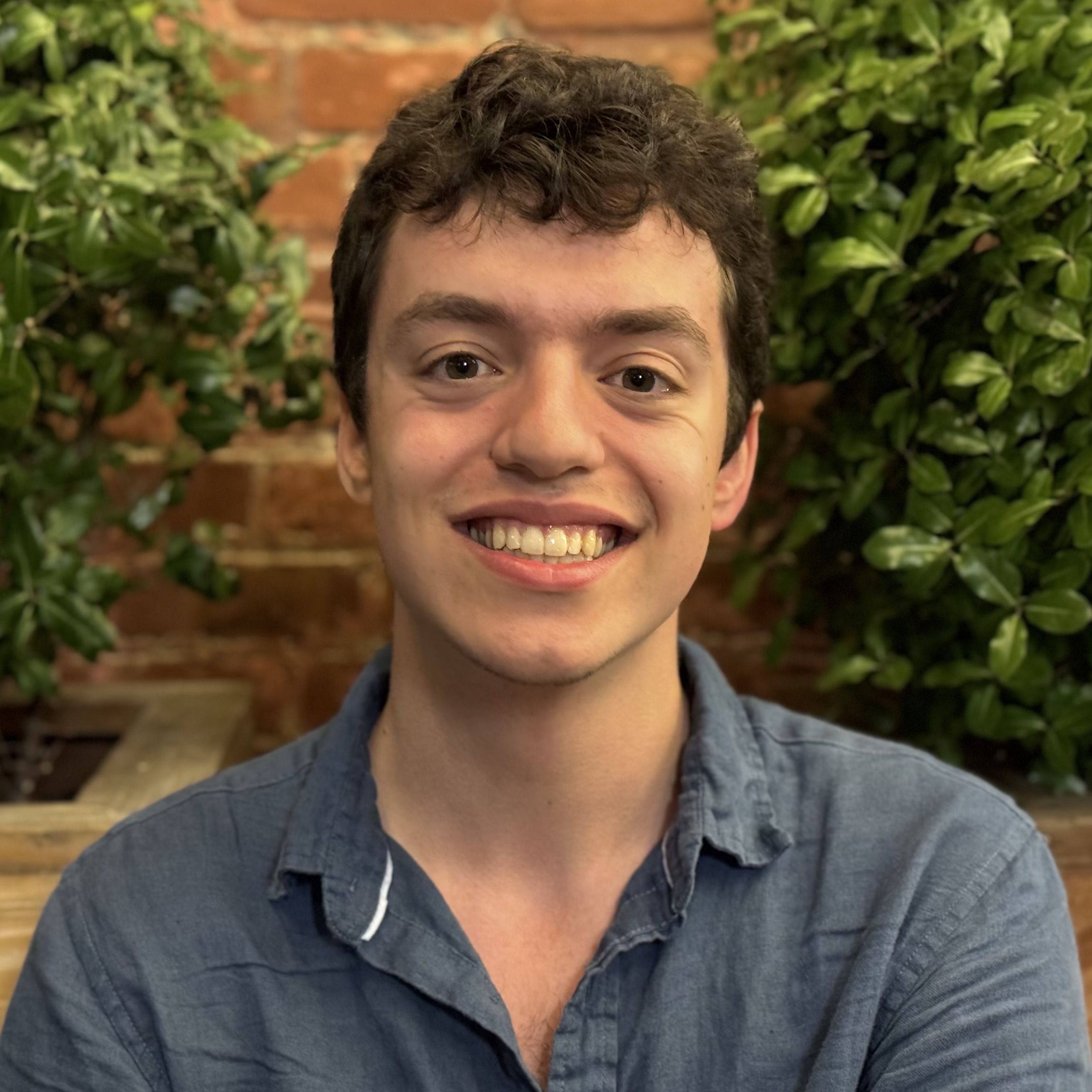 Matei standing in front of a brick wall wearing a navy blue button up shirt with one too many top buttons unbuttoned.